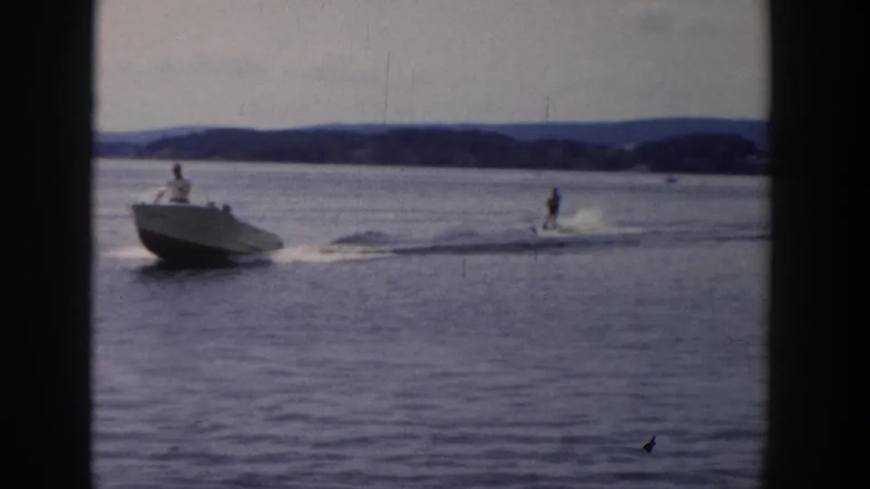 1958 Wisconsin 8mm frame showing a white motorboat towing a water skier over calm lake water near a distant shoreline.