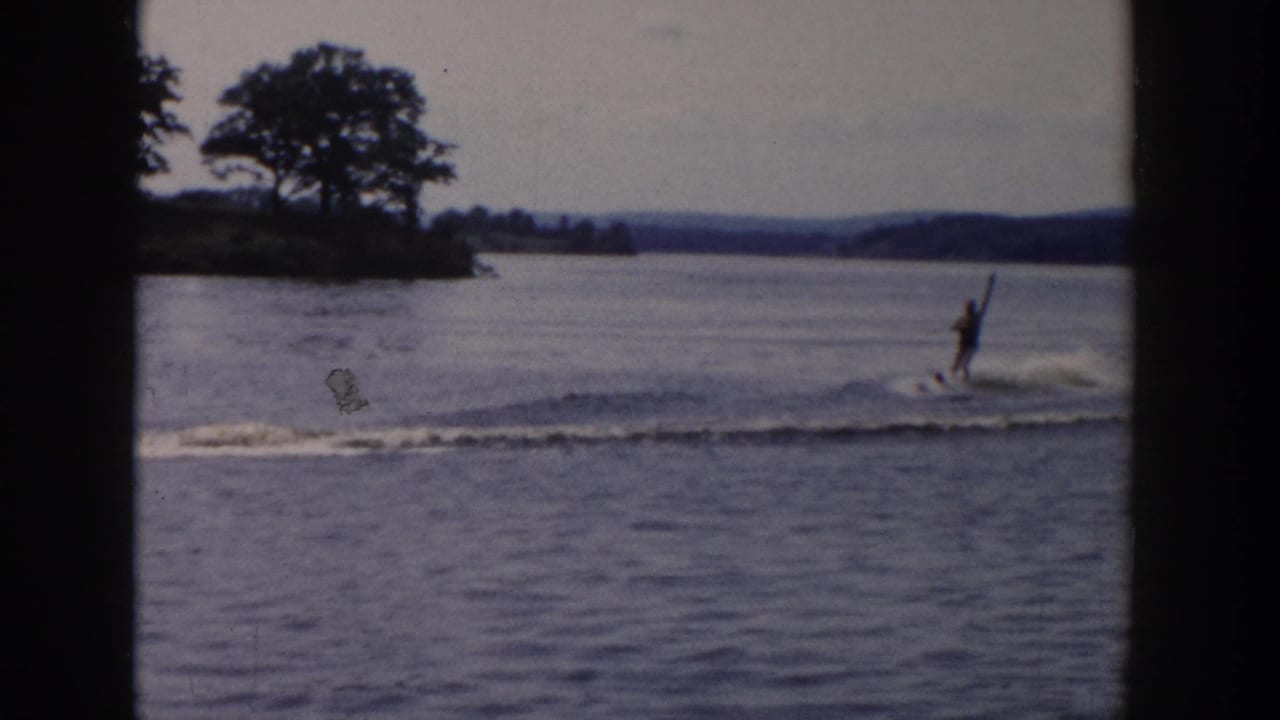 A water skier glides across the lake after the boat has moved out of view, with trees and low hills in the background.