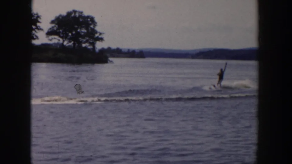 A water skier glides across the lake after the boat has moved out of view, with trees and low hills in the background.