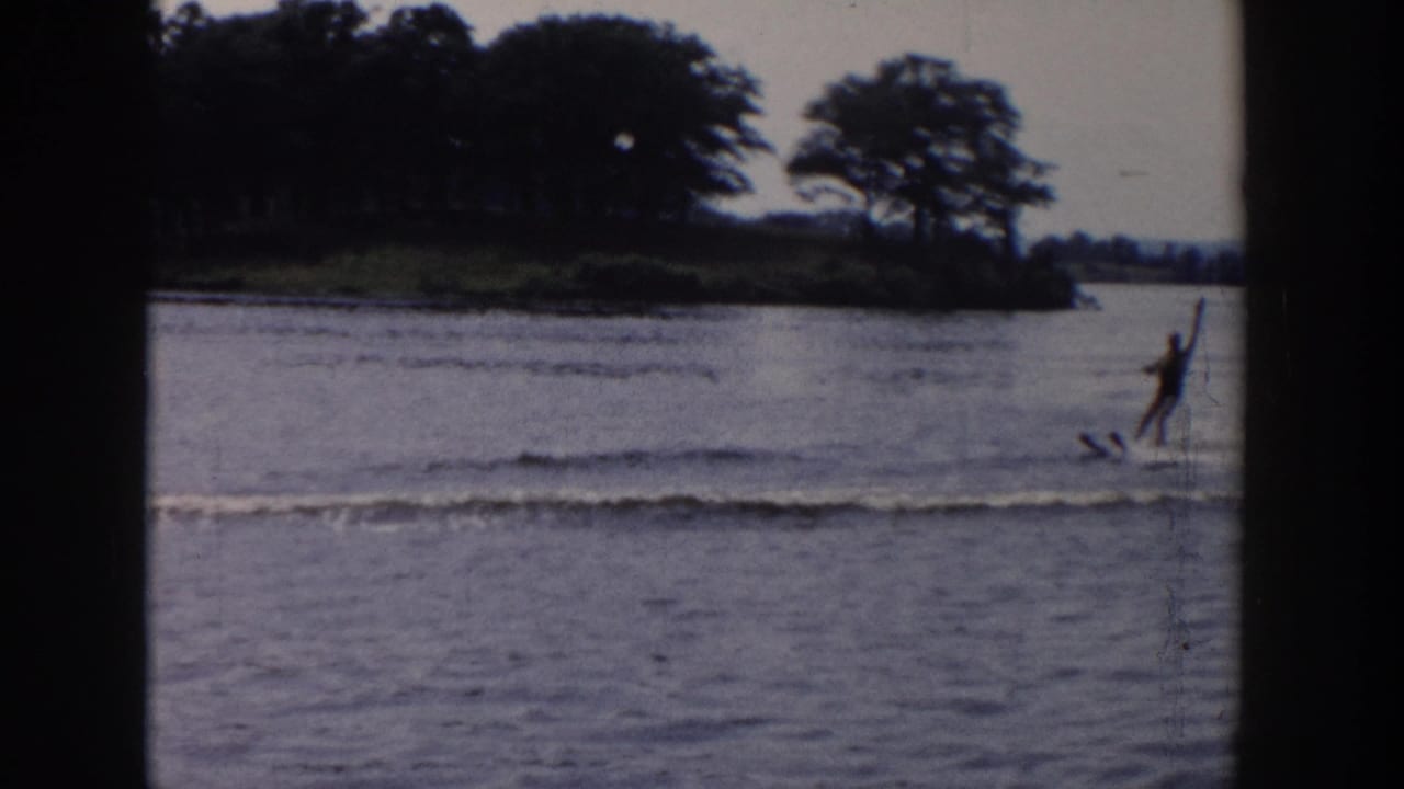 The skier continues along the lake near a wooded shoreline, with spray and muted 8mm color visible in the frame.