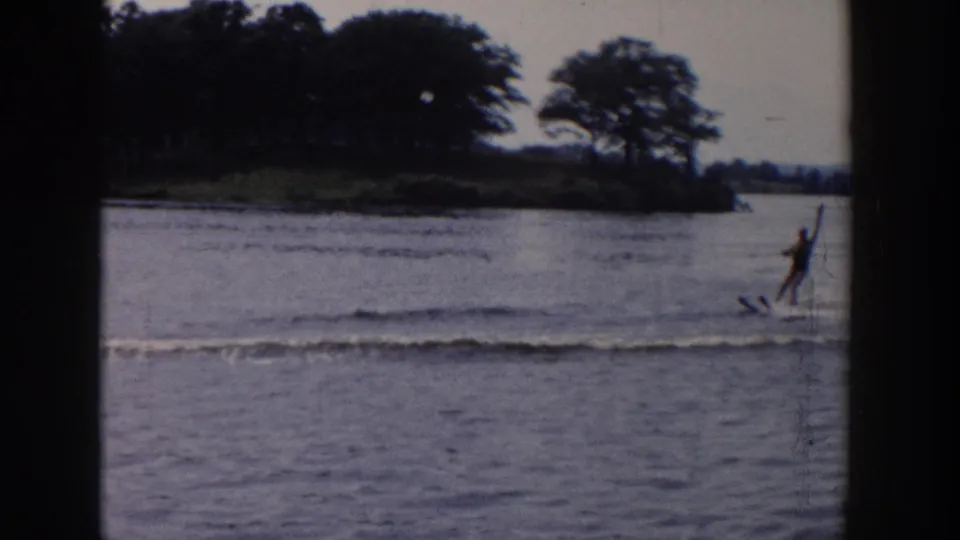 The skier continues along the lake near a wooded shoreline, with spray and muted 8mm color visible in the frame.