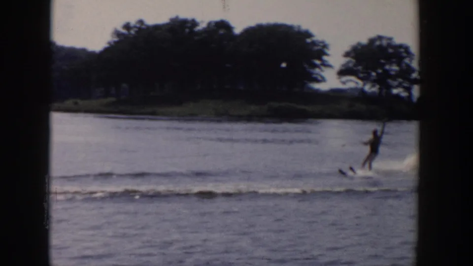 1958 Wisconsin home-movie frame showing a distant man water skiing across a lake with trees and shoreline in the background.