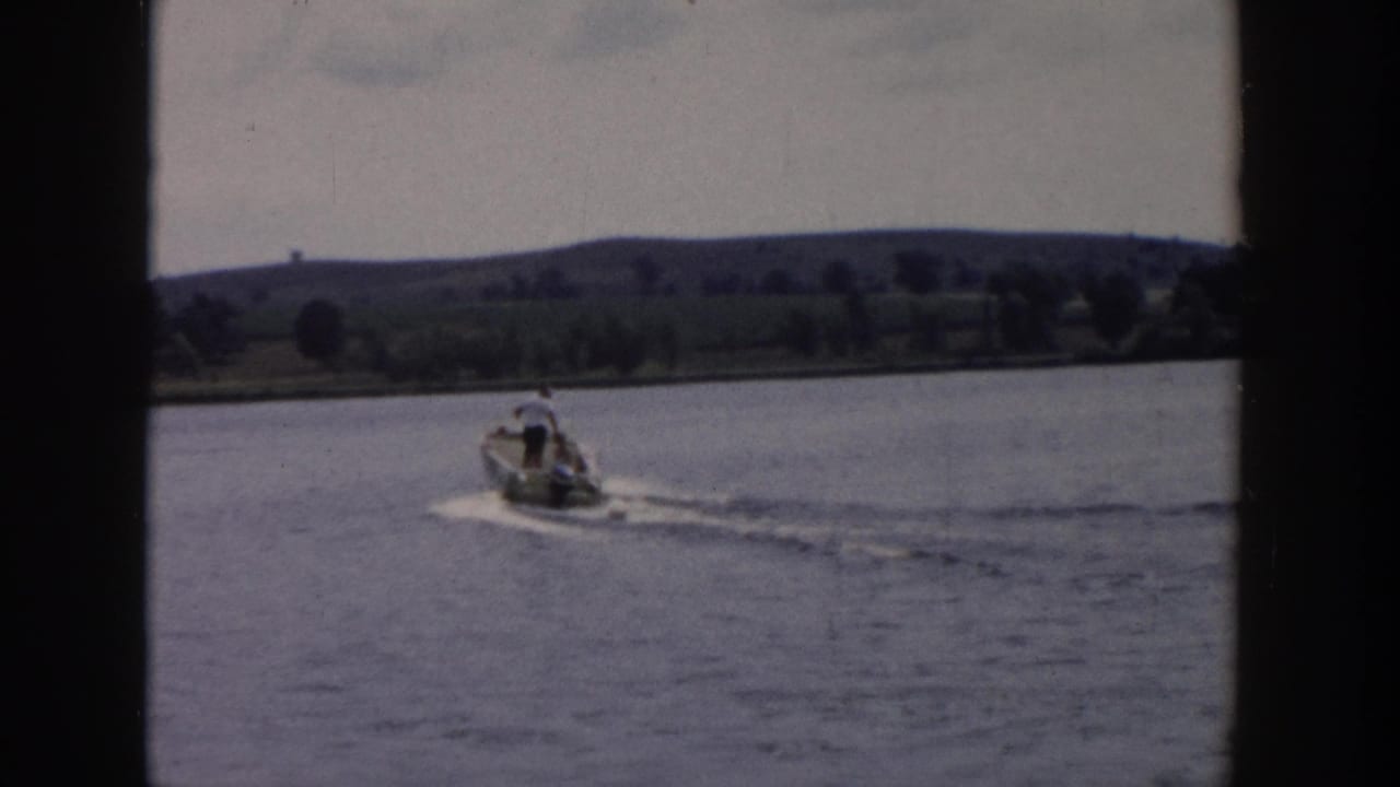 1958 Wisconsin 8mm frame showing a small motorboat crossing calm lake water with a distant tree-lined shore and low hills.
