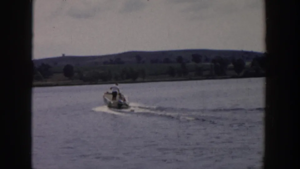 1958 Wisconsin 8mm frame showing a small motorboat crossing calm lake water with a distant tree-lined shore and low hills.