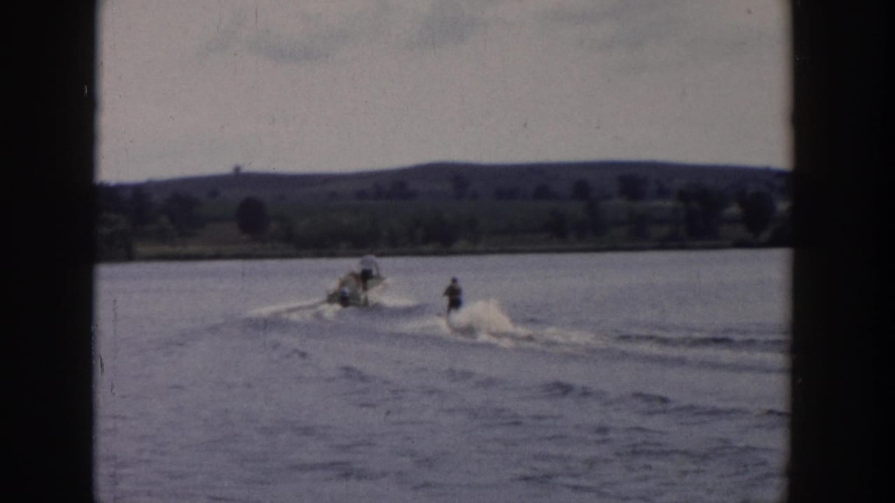 1958 Wisconsin 8mm frame with a skier trailing a motorboat on calm lake water, backed by rural hills and trees.