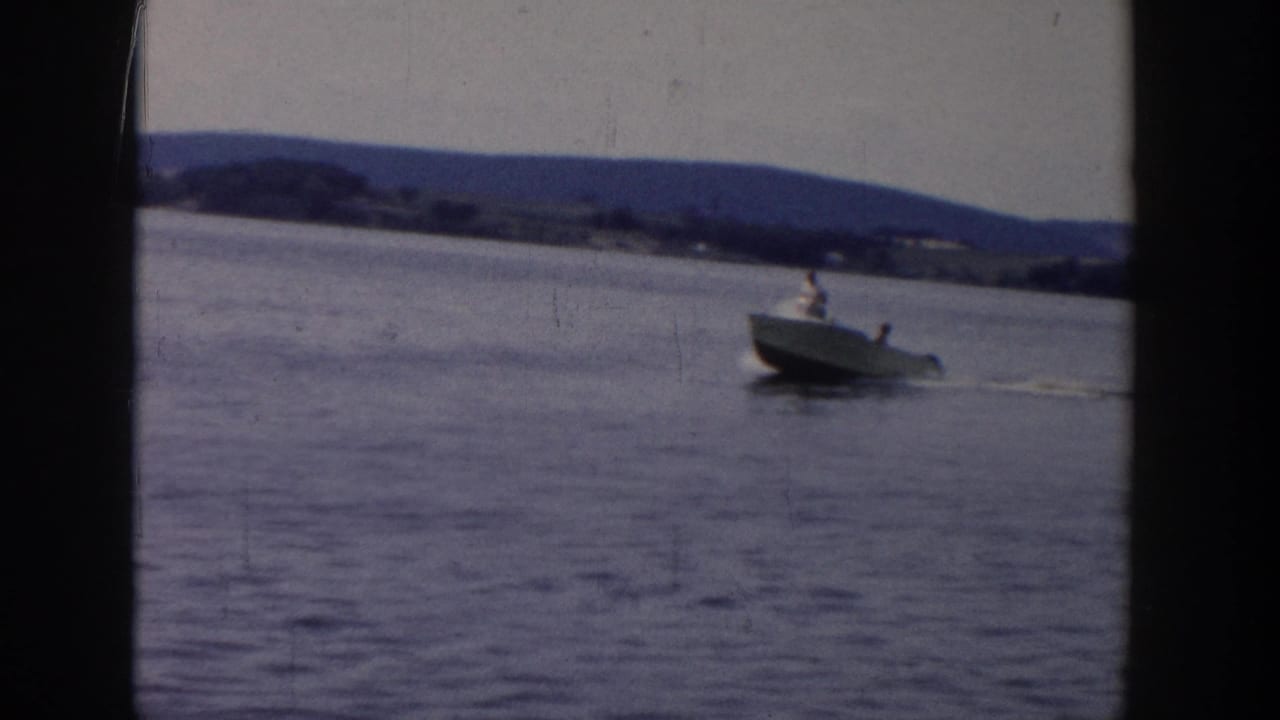 1958 Wisconsin home-movie frame showing a small motorboat on open lake water with distant hills and shoreline in view.