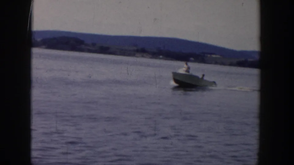 1958 Wisconsin home-movie frame showing a small motorboat on open lake water with distant hills and shoreline in view.