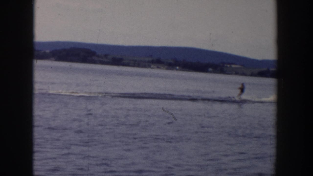 A distant water skier moves across a Wisconsin lake in 1958, with boat wake and low shoreline hills behind.