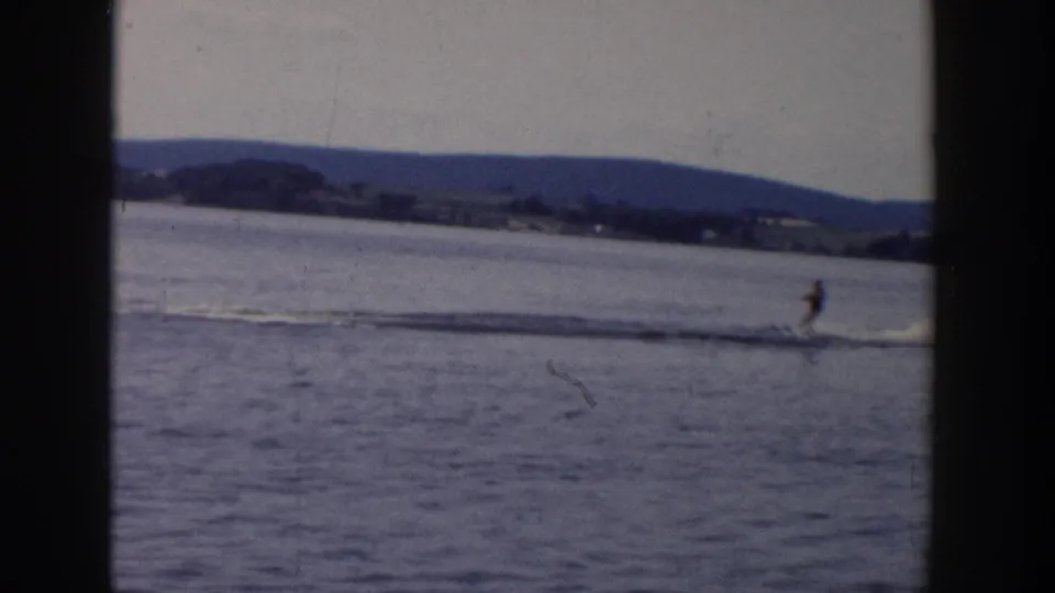 A distant water skier moves across a Wisconsin lake in 1958, with boat wake and low shoreline hills behind.