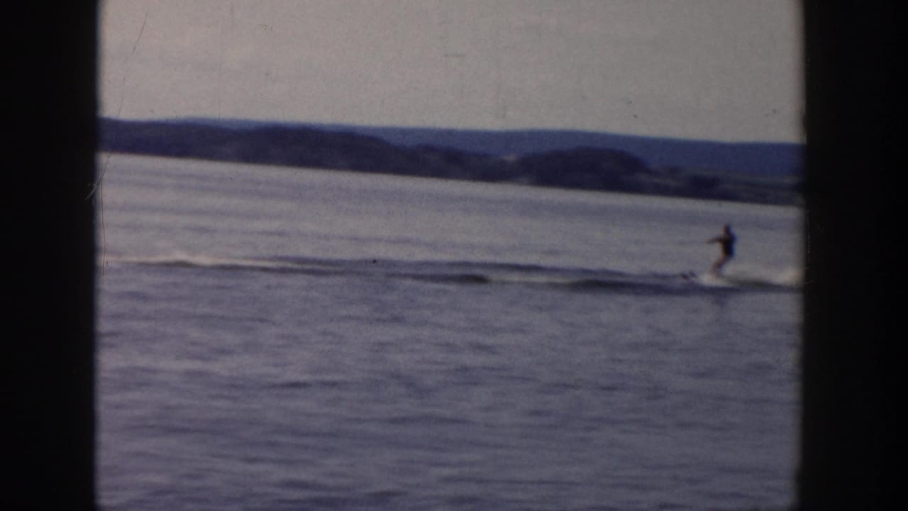 1958 Wisconsin home-movie frame of a water skier gliding across calm water with hills and shoreline in the background.