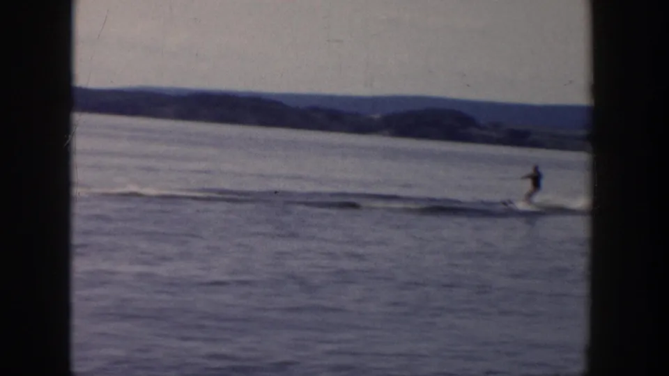 1958 Wisconsin home-movie frame of a water skier gliding across calm water with hills and shoreline in the background.