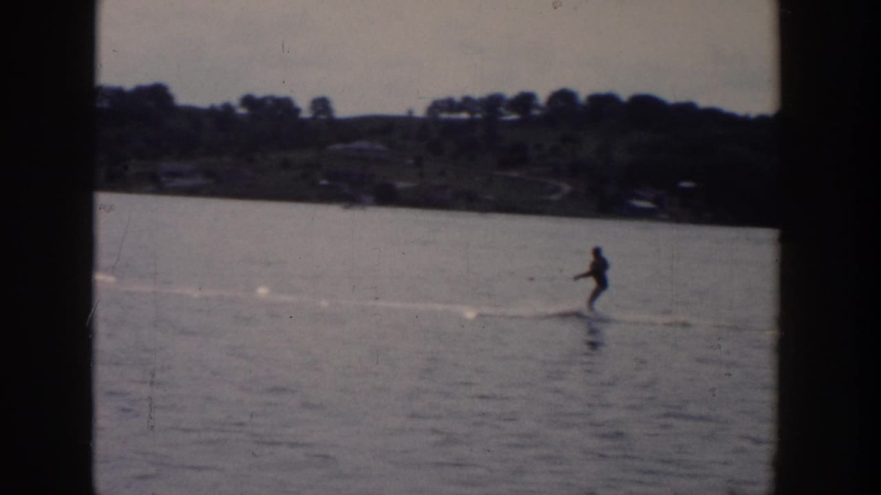 A distant skier rides over calm Wisconsin lake water in 1958, framed by a wooded shoreline and soft 8mm exposure.