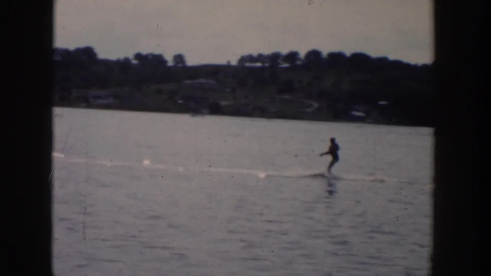 A distant skier rides over calm Wisconsin lake water in 1958, framed by a wooded shoreline and soft 8mm exposure.