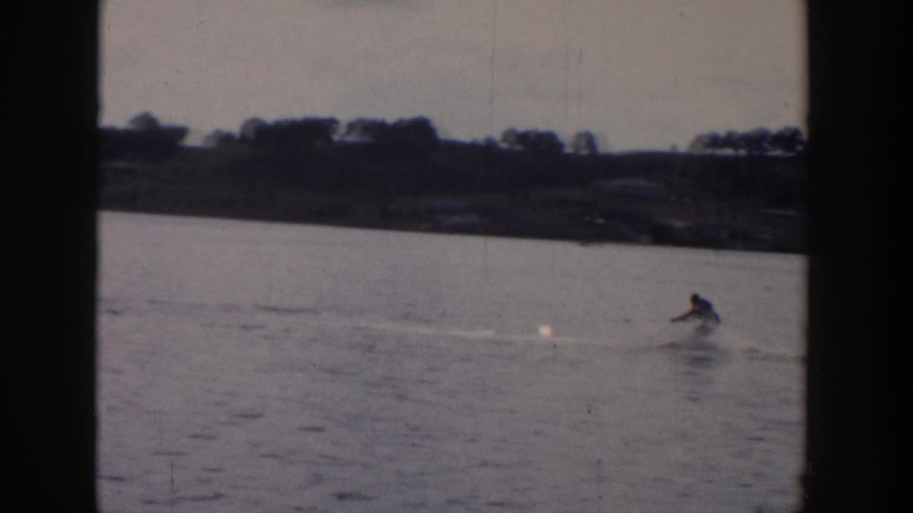 1958 Wisconsin 8mm frame showing a distant person water skiing across a calm lake under an overcast sky.