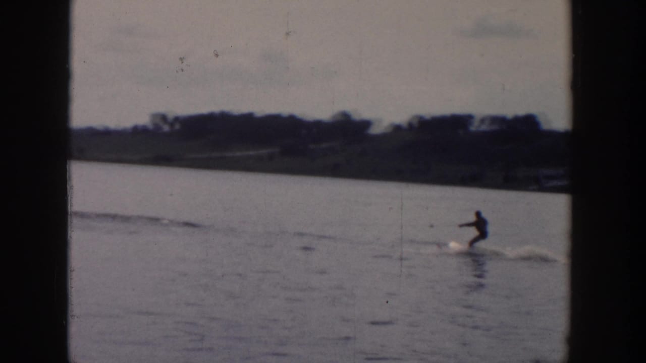 A distant skier in dark clothing glides across a Wisconsin lake, with wake lines and shoreline visible behind.