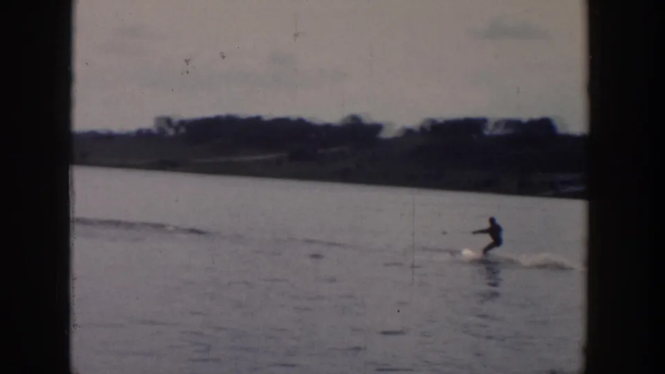 A distant skier in dark clothing glides across a Wisconsin lake, with wake lines and shoreline visible behind.