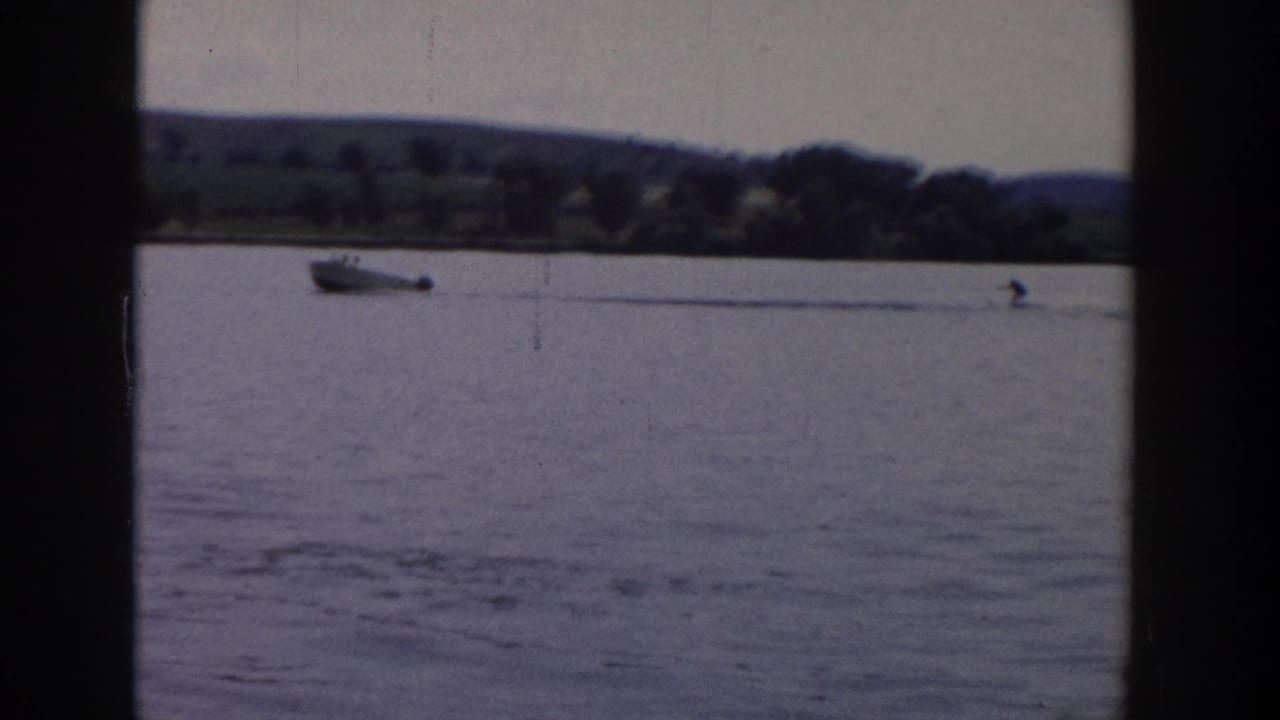 Motorboat and distant skier crossing open water near a tree-lined shore.