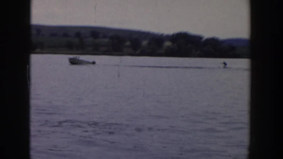 A 1958 Wisconsin home-movie frame captures the boat and water skier farther across the lagoon, with trees and low hills along the shoreline.