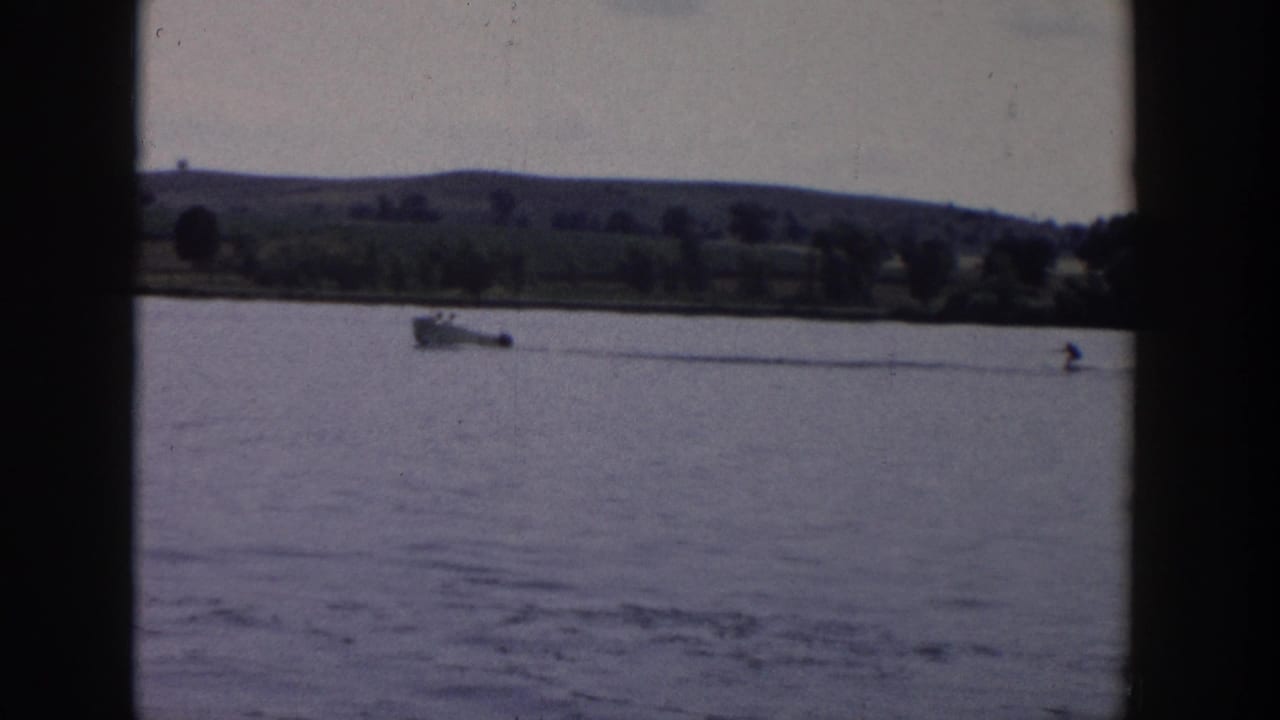 Vintage 8mm footage from Wisconsin in 1958 shows the motorboat towing a distant skier across calm water, framed by a broad shoreline and hills.