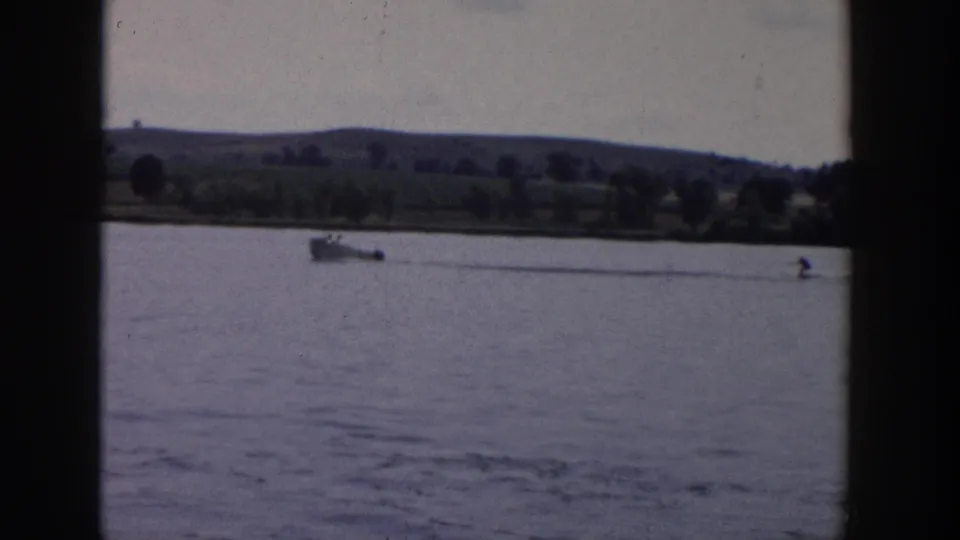 Vintage 8mm footage from Wisconsin in 1958 shows the motorboat towing a distant skier across calm water, framed by a broad shoreline and hills.