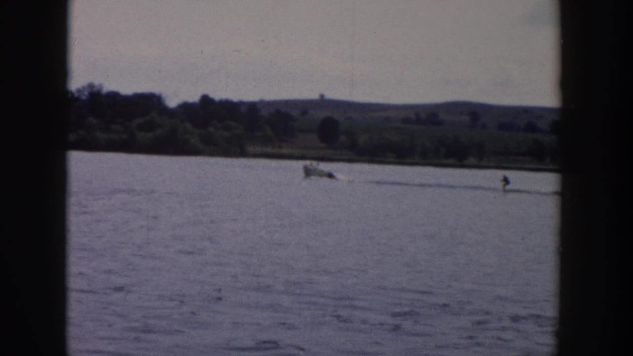 Distant motorboat pulling a water skier across a lake near a wooded shoreline.