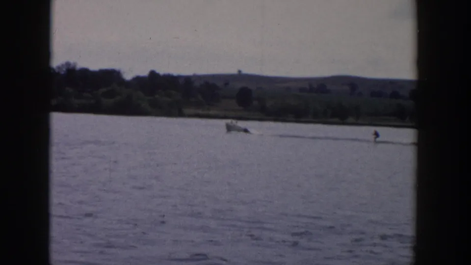 1958 Wisconsin 8mm frame showing a small boat towing a water skier across open lake water with low hills and trees beyond.