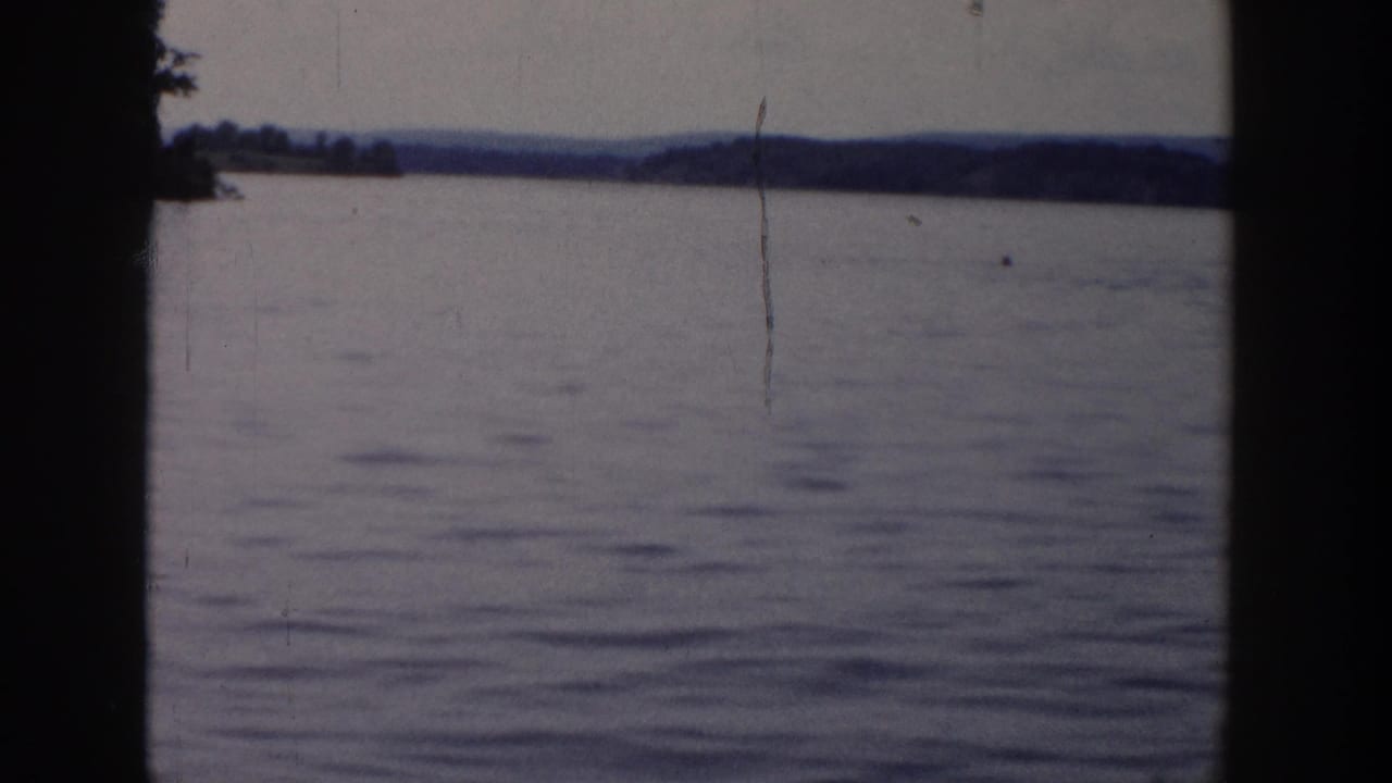 1958 Wisconsin home-movie frame of open lake water and distant wooded hills, marked by grain, scratches, and dark 8mm frame edges.