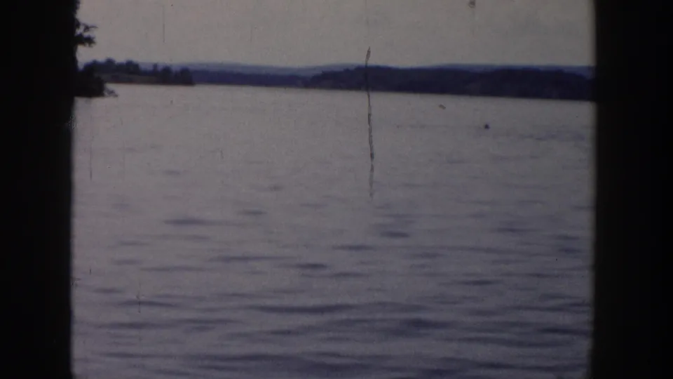 1958 Wisconsin home-movie frame of open lake water and distant wooded hills, marked by grain, scratches, and dark 8mm frame edges.