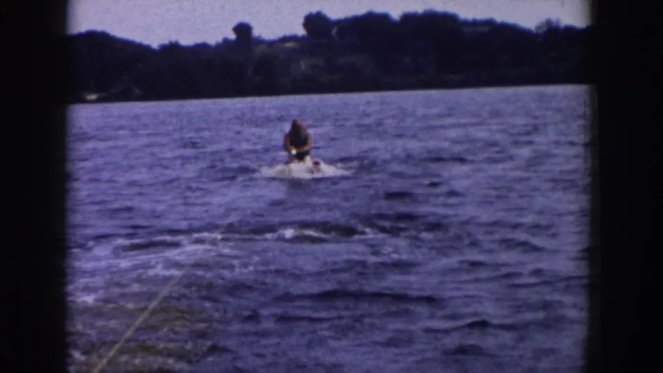 1958 Wisconsin home-movie frame with a skier rising behind a tow rope on a lake, with trees and open sky in the background.