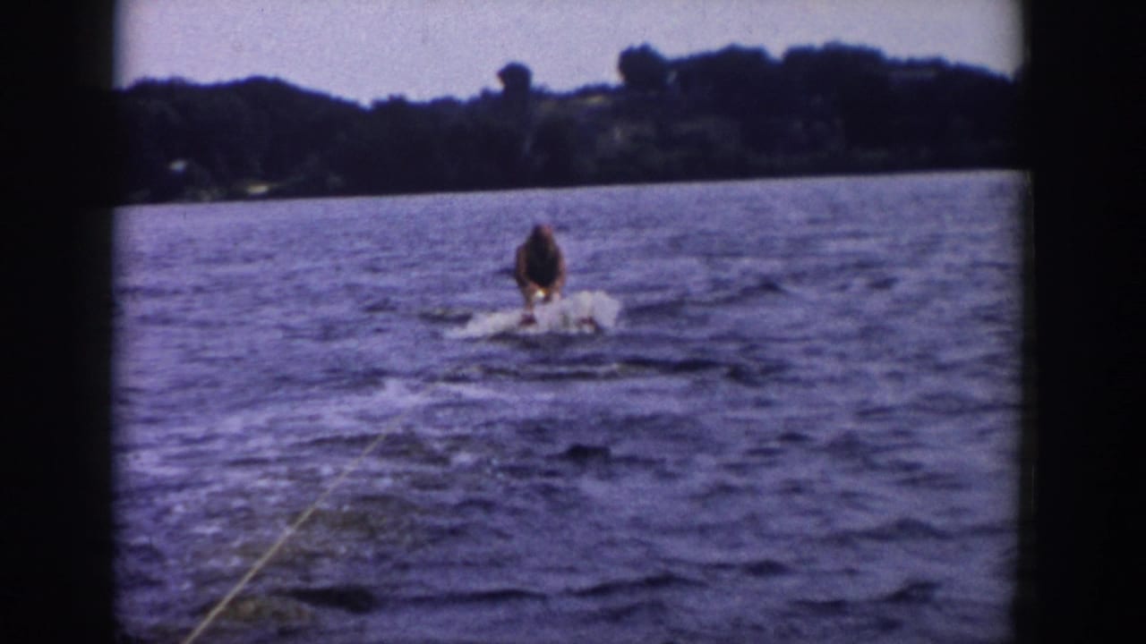 1958 Wisconsin 8mm still of a water skier gaining balance, with spray, tow rope, rippled lake water, and wooded shore visible.