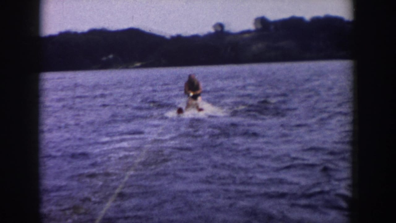 1958 Wisconsin home-movie frame showing the skier continuing on the lake, with tow rope, wake, blue water, and distant trees.