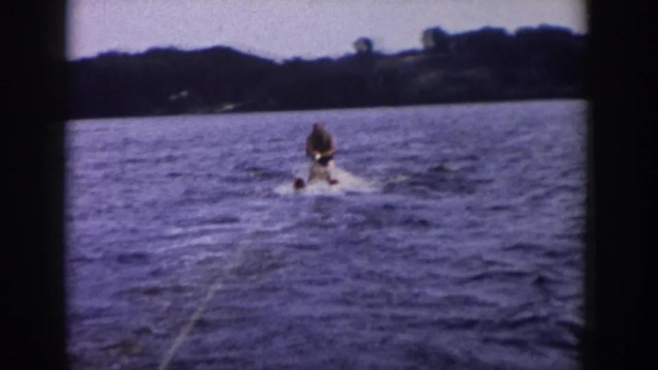 1958 Wisconsin home-movie frame showing the skier continuing on the lake, with tow rope, wake, blue water, and distant trees.