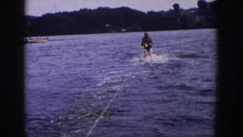 1958 Wisconsin home-movie frame showing a man water skiing across a lake, seen from behind a tow rope with trees on the far shore.