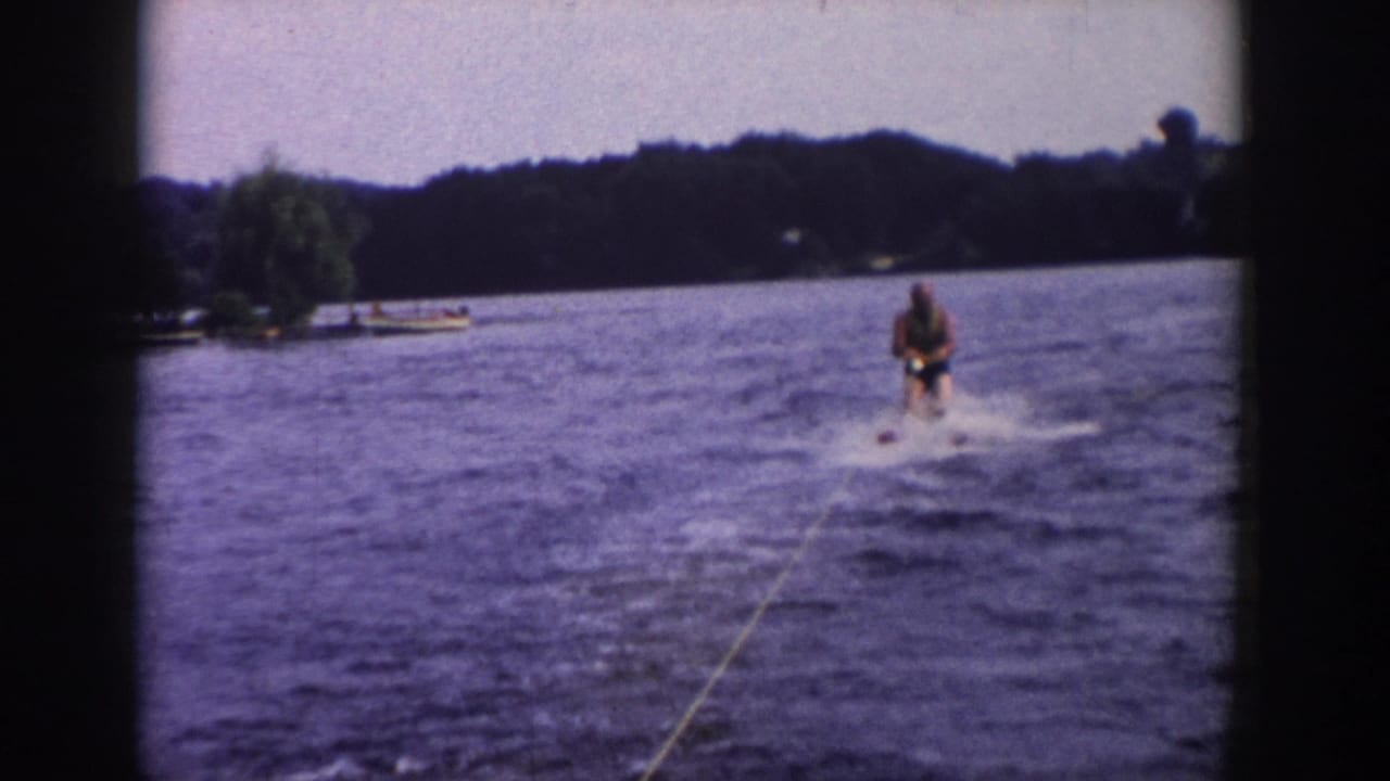 Water skier rides over the boat wake with a tree-lined shore behind him.