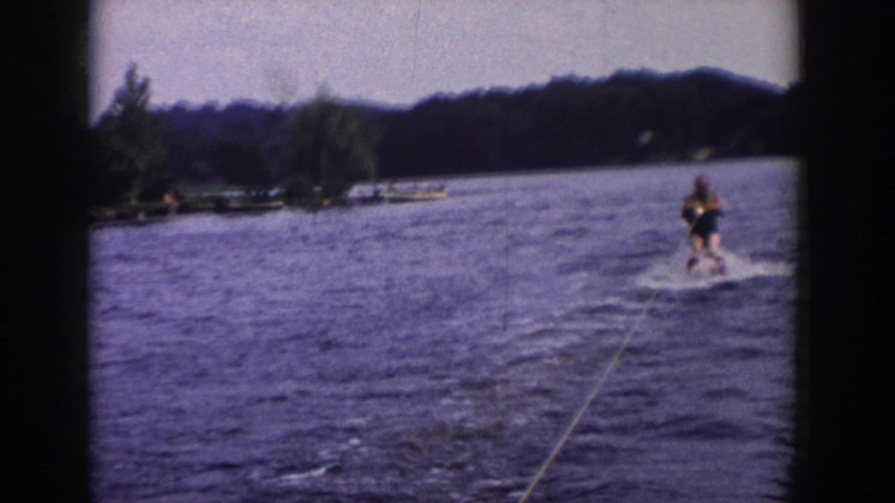 The 1958 Wisconsin lake scene shows a distant man water skiing behind the tow boat, with shoreline trees and dock or boat shapes nearby.