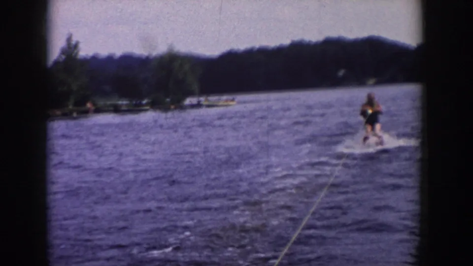 The 1958 Wisconsin lake scene shows a distant man water skiing behind the tow boat, with shoreline trees and dock or boat shapes nearby.