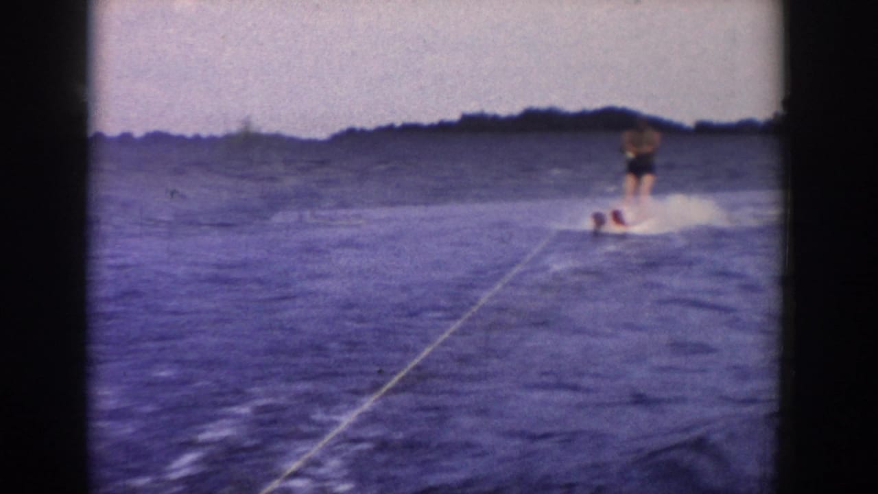 A man water skis at the right side of the frame on a Wisconsin lake, leaning across the wake with the tow rope stretching toward the camera.