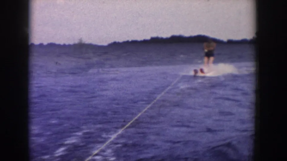 A man water skis at the right side of the frame on a Wisconsin lake, leaning across the wake with the tow rope stretching toward the camera.