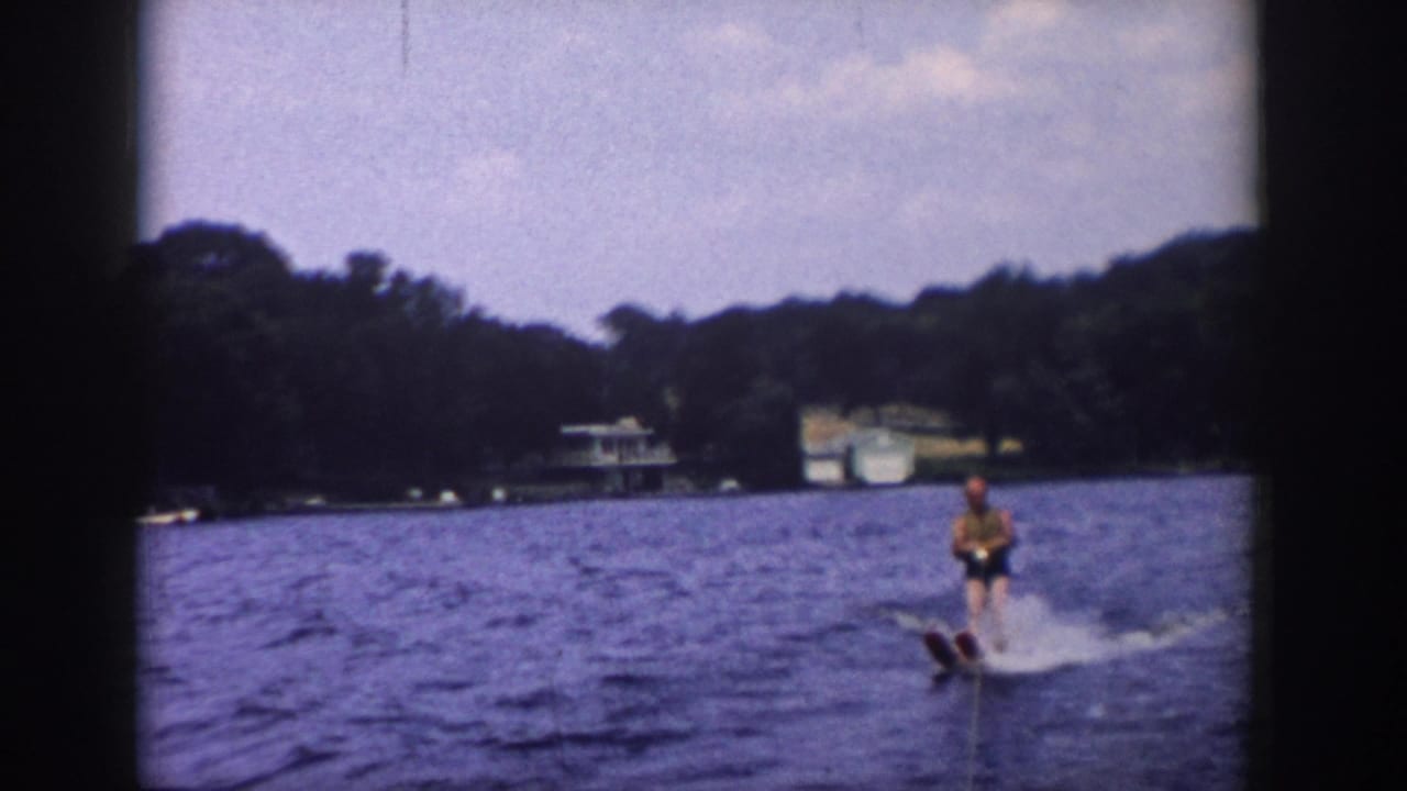 Wider 1958 Wisconsin lake view with a water skier, tow rope, wooded shoreline, and distant lakeside structures.
