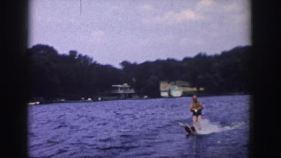 Wider 1958 Wisconsin lake view with a water skier, tow rope, wooded shoreline, and distant lakeside structures.