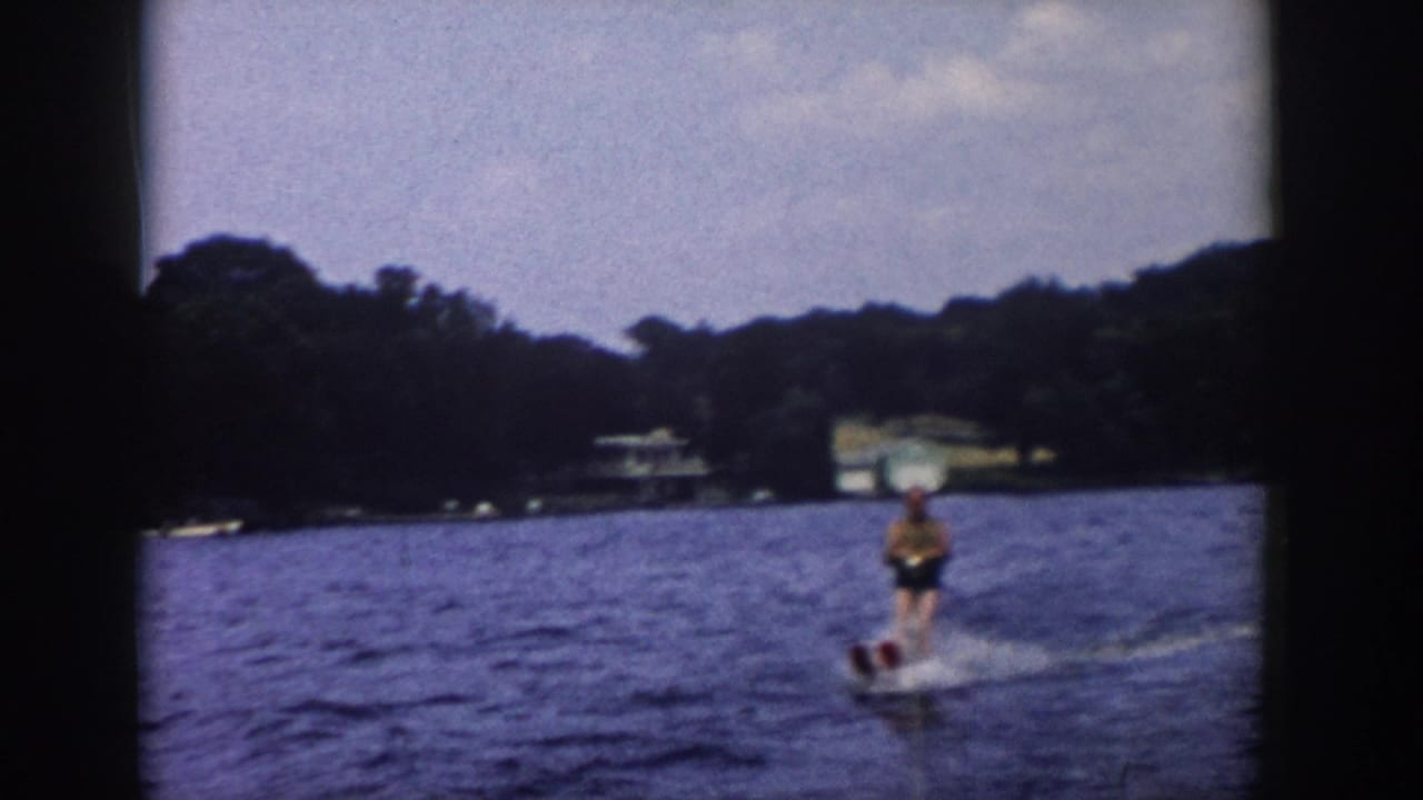 Water skier moves across a lake with forested shoreline and distant buildings behind.