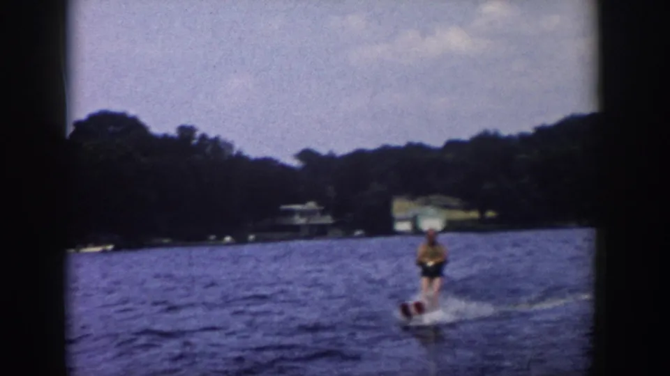 1958 Wisconsin 8mm frame showing a water skier on a lake, with white spray, wooded shoreline, and distant lakeside buildings.