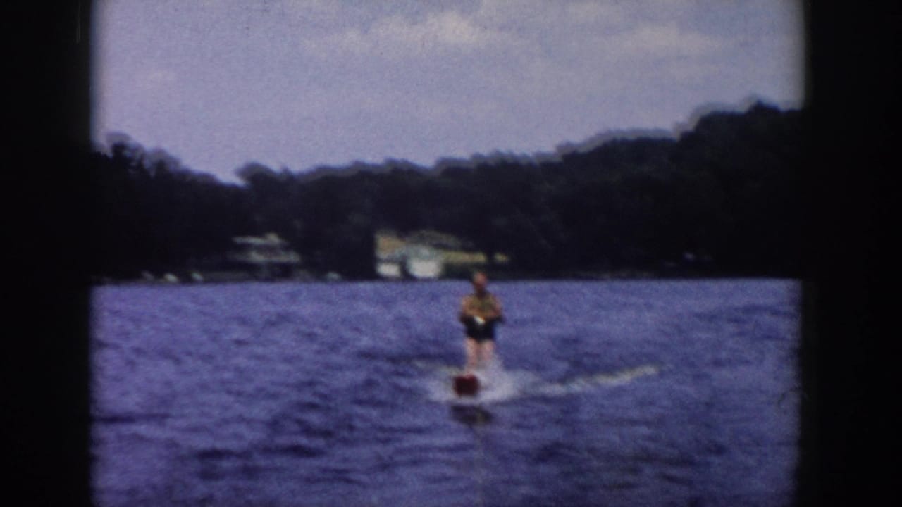 1958 Wisconsin home-movie frame of a water skier moving away across blue lake water, with forested shore and distant buildings behind.