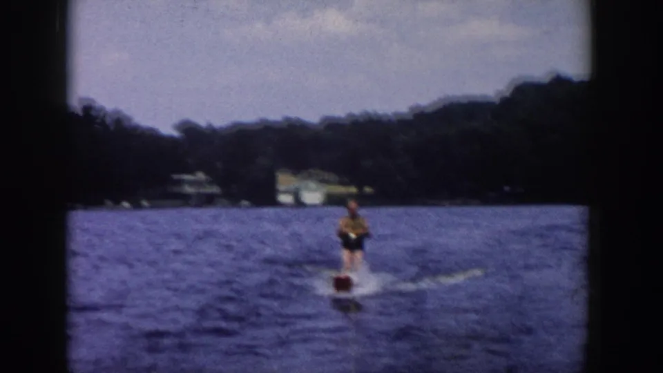 1958 Wisconsin home-movie frame of a water skier moving away across blue lake water, with forested shore and distant buildings behind.