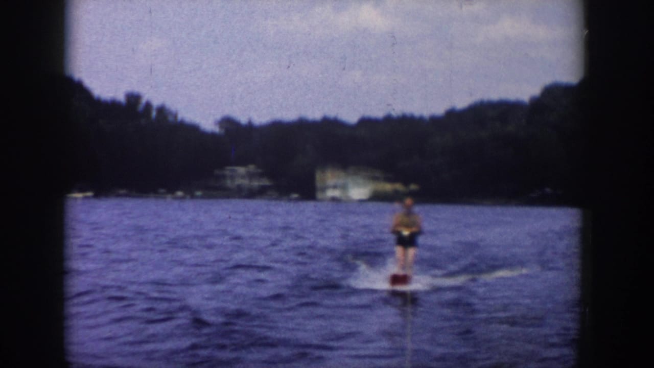 In 1958 Wisconsin home-movie footage, an adult water skier trails behind a boat on blue water with trees and a distant house beyond.