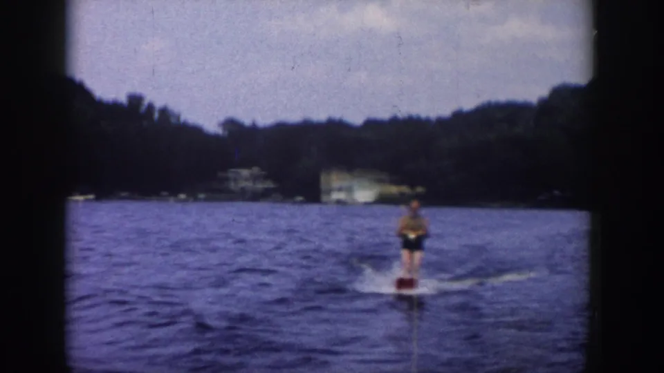 In 1958 Wisconsin home-movie footage, an adult water skier trails behind a boat on blue water with trees and a distant house beyond.