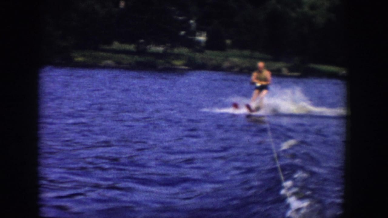 The skier continues across the blue water in Wisconsin, with the tow rope, wake, spray, and dark shoreline visible in the frame.