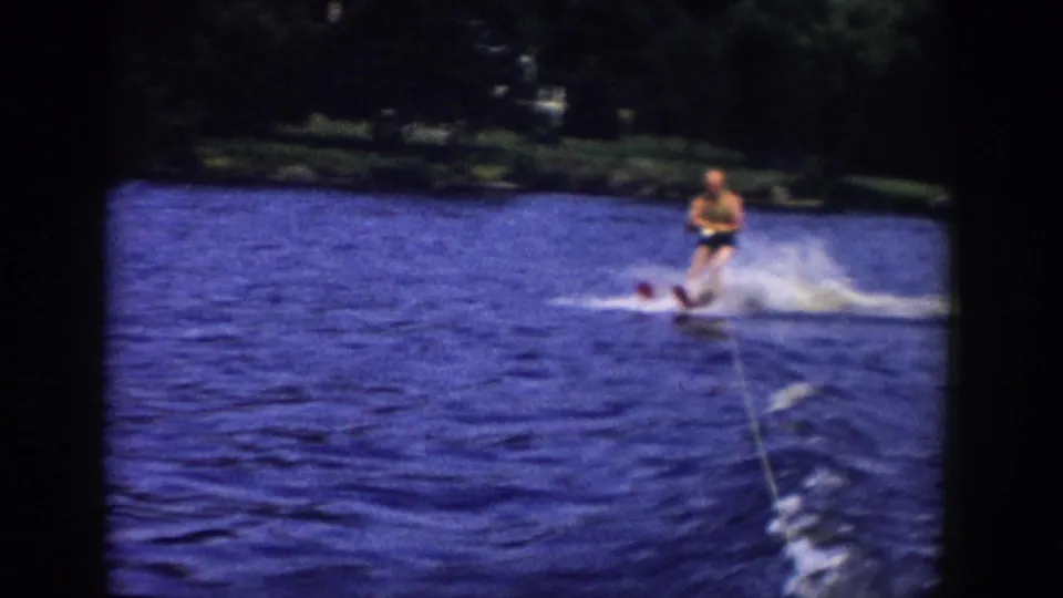 The skier continues across the blue water in Wisconsin, with the tow rope, wake, spray, and dark shoreline visible in the frame.
