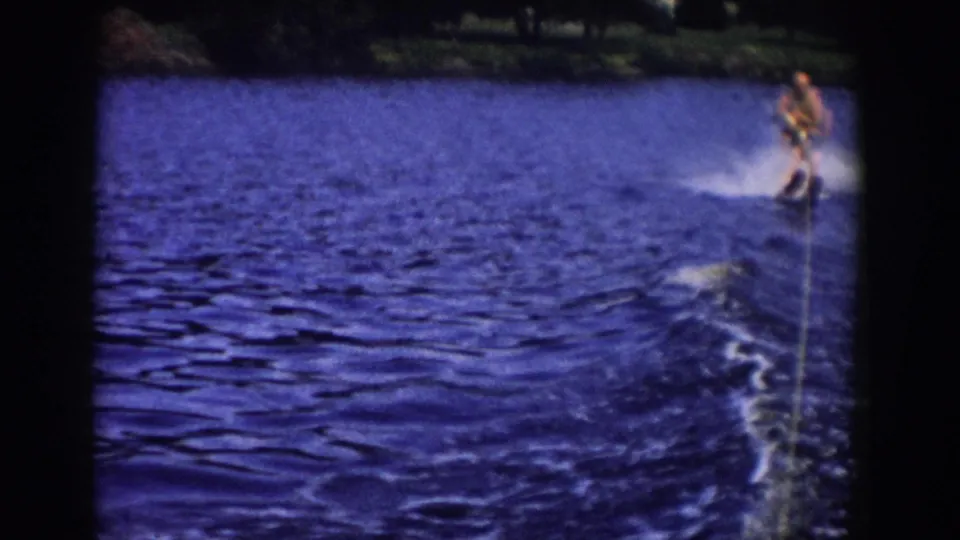 1958 Wisconsin home-movie frame showing a single water skier behind a boat on a lake, with wake, spray, and shoreline in the distance.