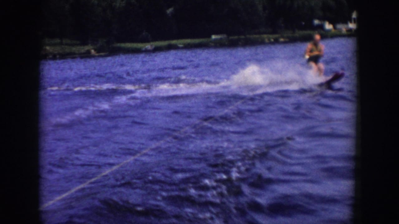 Home-movie frame of one person water skiing behind a boat, with the rope visible at lower left and wooded Wisconsin shoreline beyond.